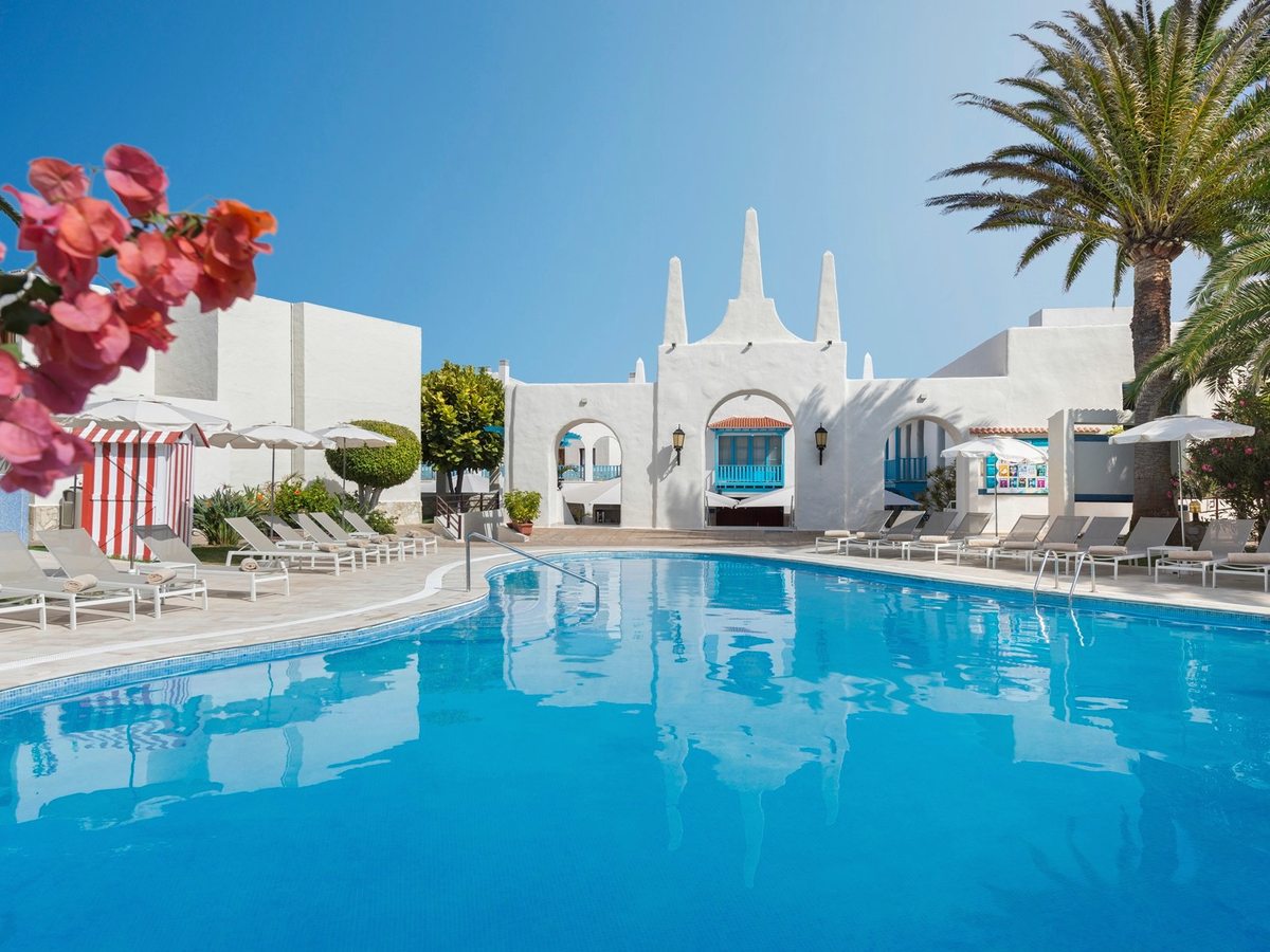 A large pool reflecting a whitewashed Canary-style building with spired gables and a blue balcony, with loungers, parasols and bougainvillea around the deck.