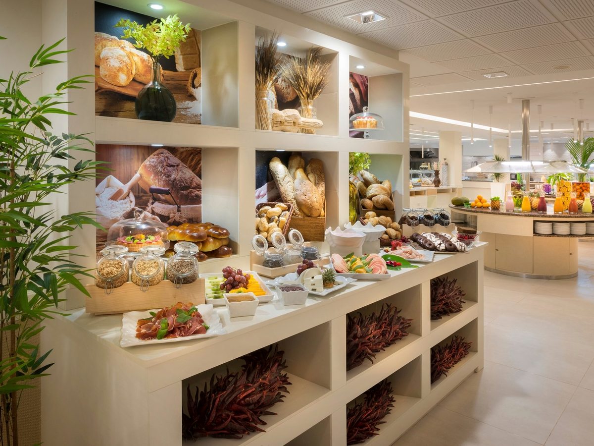 A buffet island stacked with breads, pastries, fresh fruit and cold meats in front of tall windows.