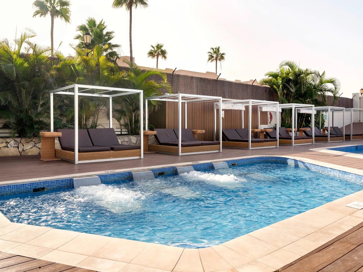 A row of four-poster cabana daybeds along the deck of a pool fed by water jets, screened by palms and a reed fence.