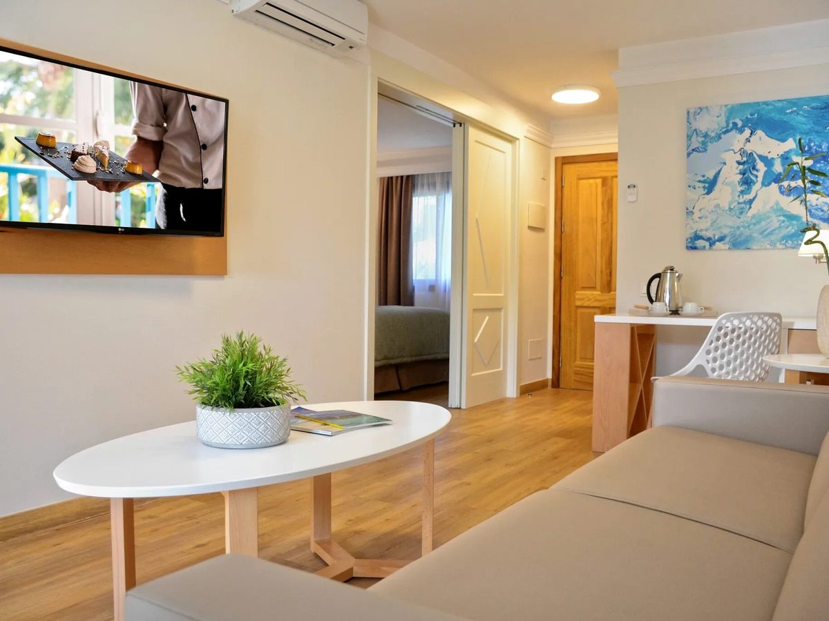 A suite lounge with a sofa, a round coffee table, a wall-mounted TV and a tea and coffee station, with an open doorway through to a bedroom.