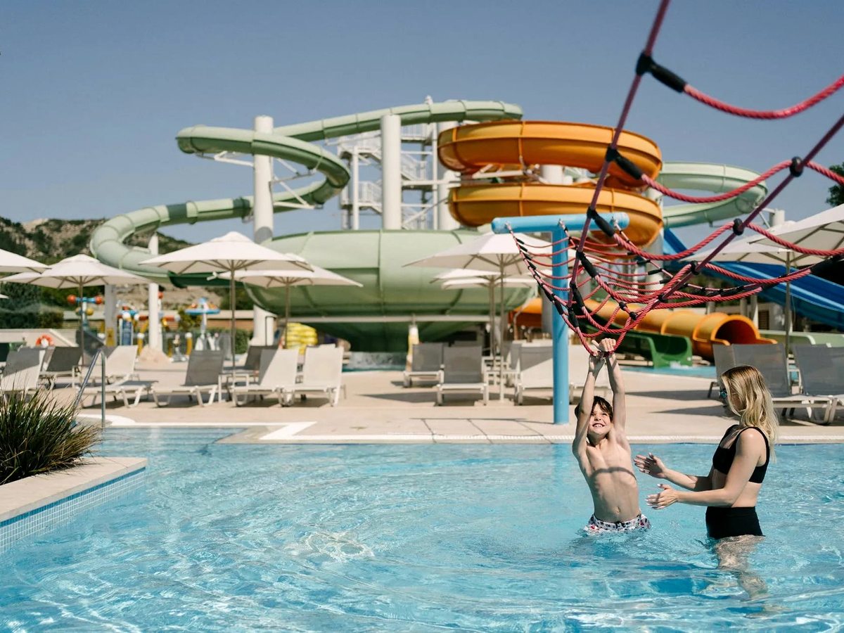 A boy swings across a rope net over a shallow pool, with the tall green and orange slide tower rising behind under a clear sky.