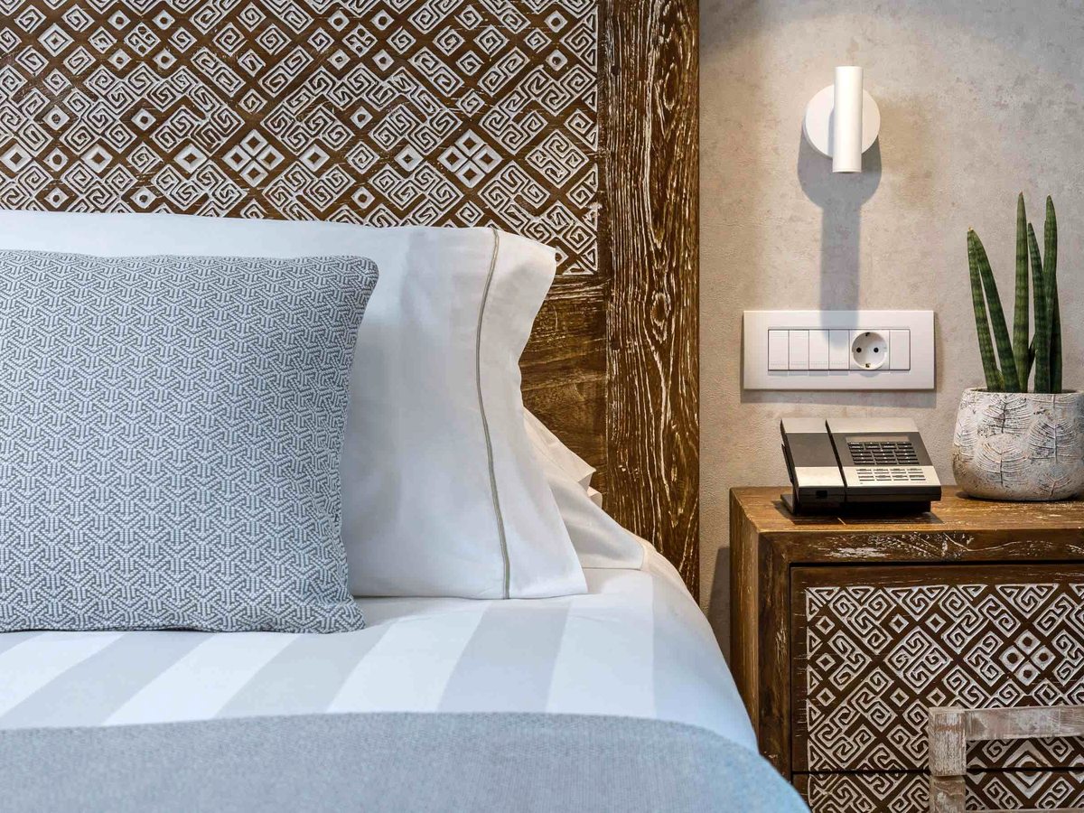 A made bed with a blue-grey woven cushion and white pillows against a carved wooden headboard patterned with Greek-key motifs, and a matching bedside table with a plant.