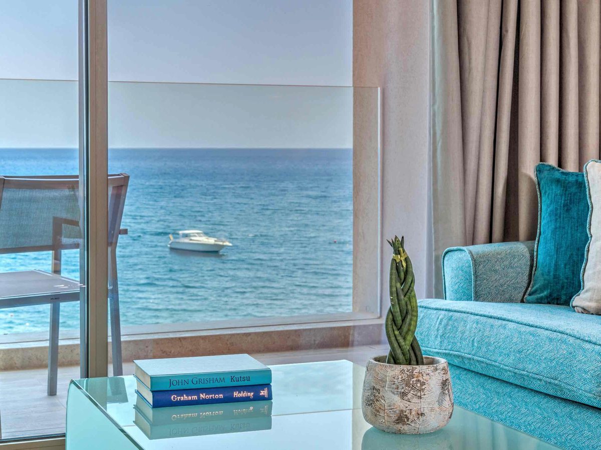 A turquoise sofa and glass coffee table by floor-to-ceiling balcony windows looking onto the sea, with a small boat on the water.