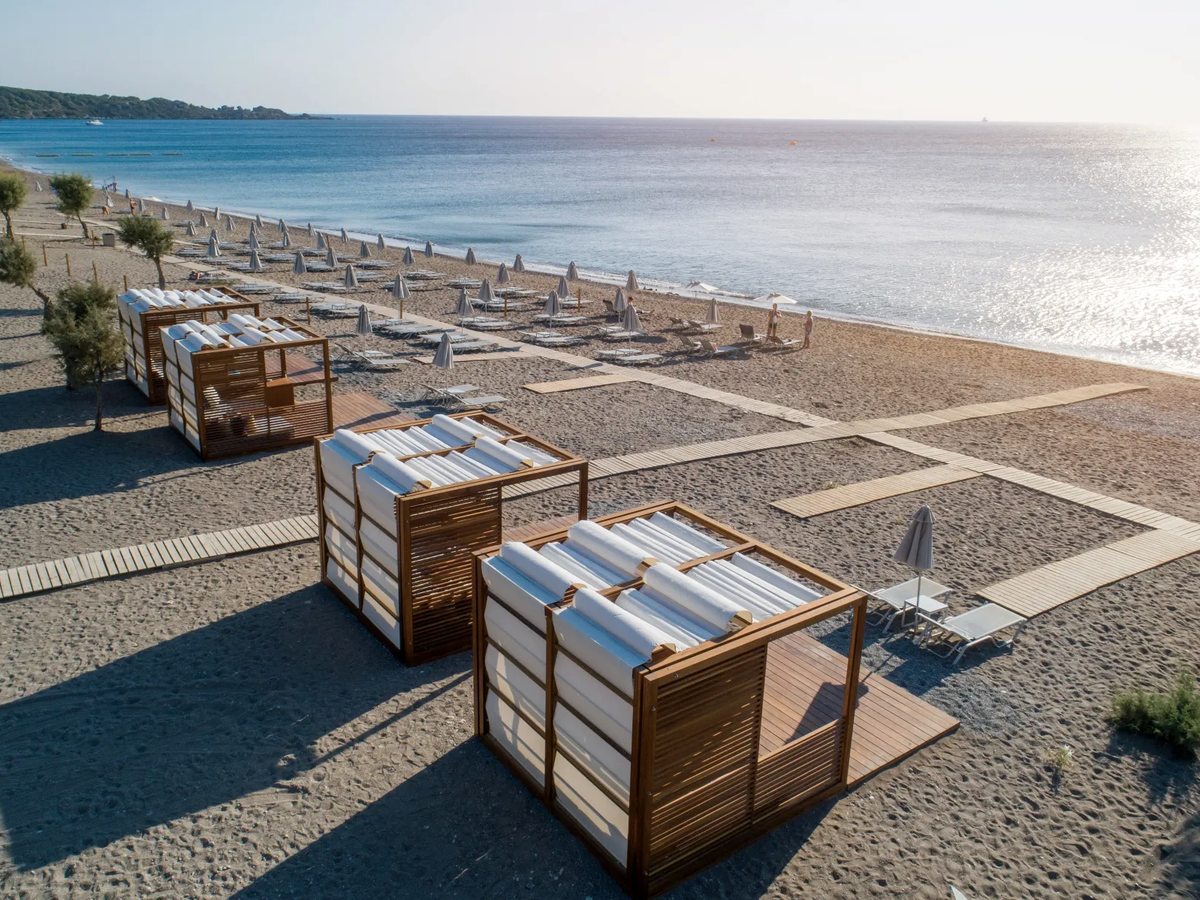 Wooden slatted beach cabanas with rolled sunbeds stand on the sand above rows of grey parasols along the shore, the calm sea to the right.