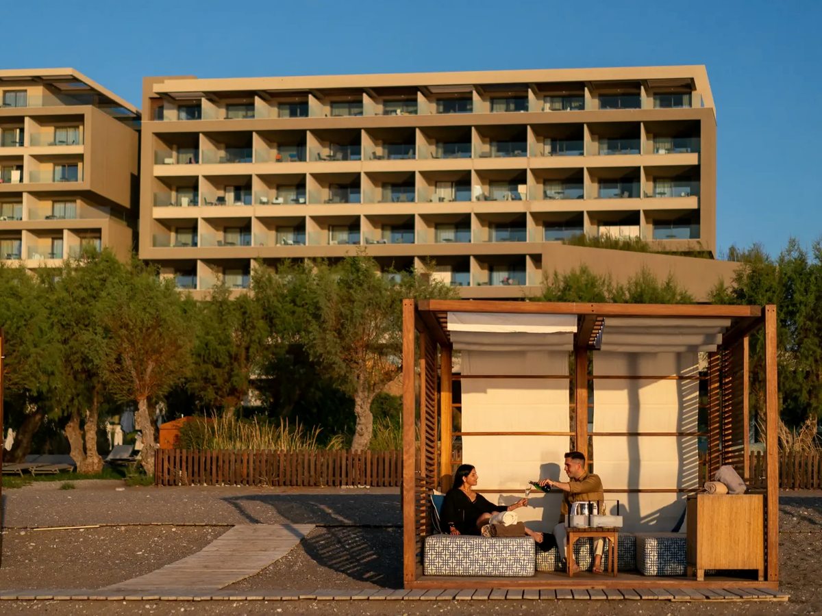 A multi-storey resort block of balconied rooms rises behind an ochre timber garden pergola where two people sit, under a clear sky.