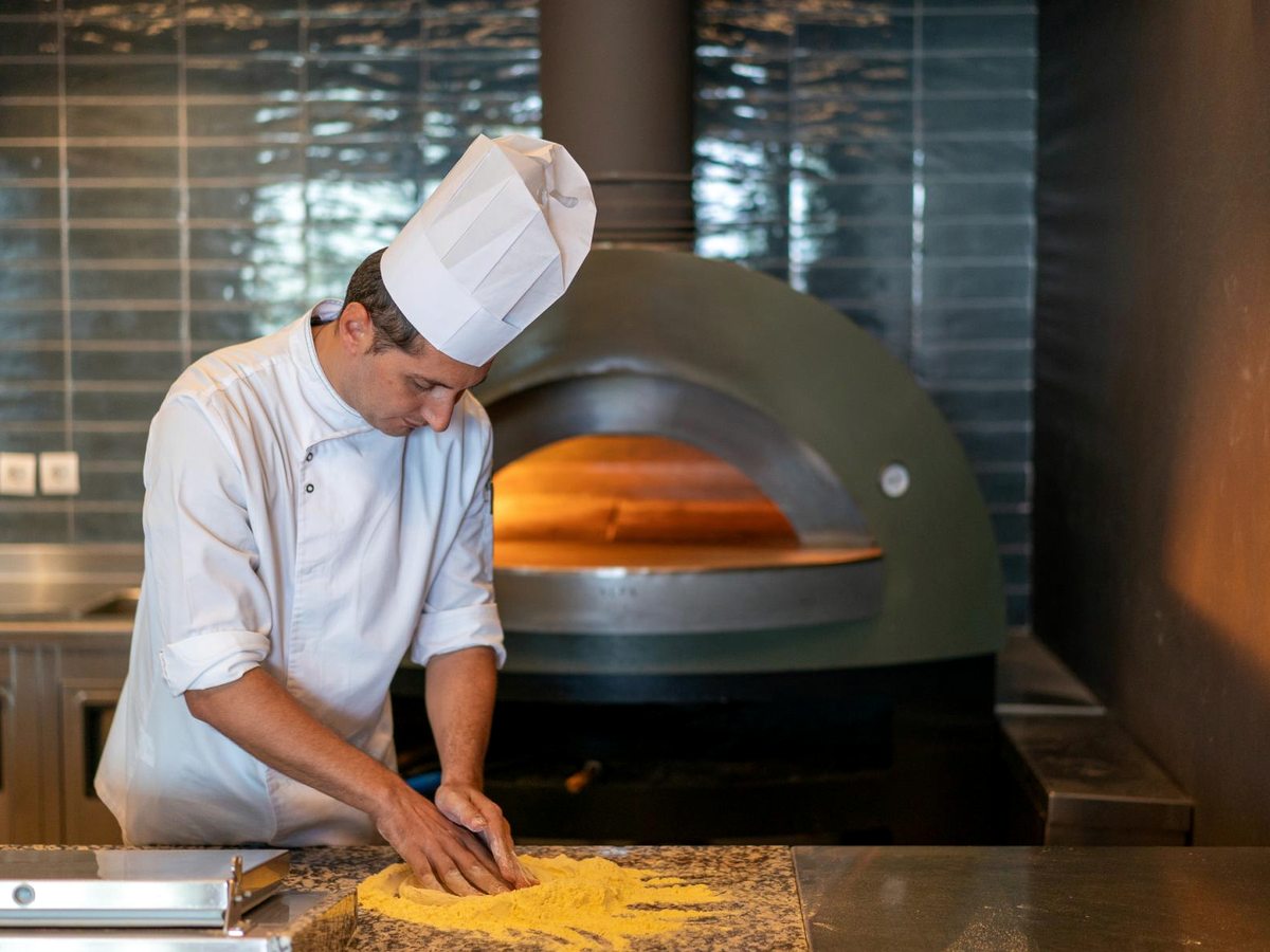 A chef in whites shaping pizza dough on a floured counter beside a green domed wood-fired oven glowing inside.