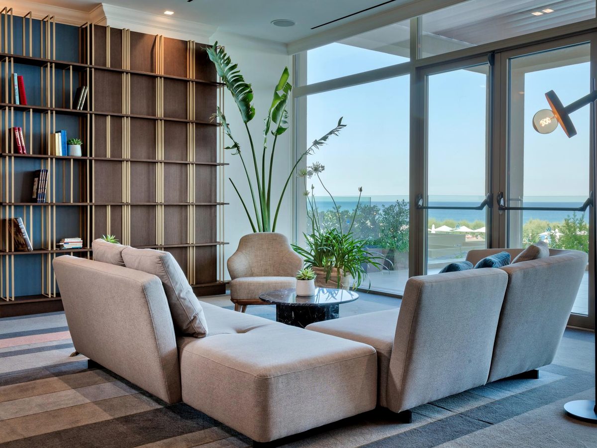 An indoor lounge with cream sofas and armchairs, a full-height bookcase and floor-to-ceiling windows looking out to the sea.