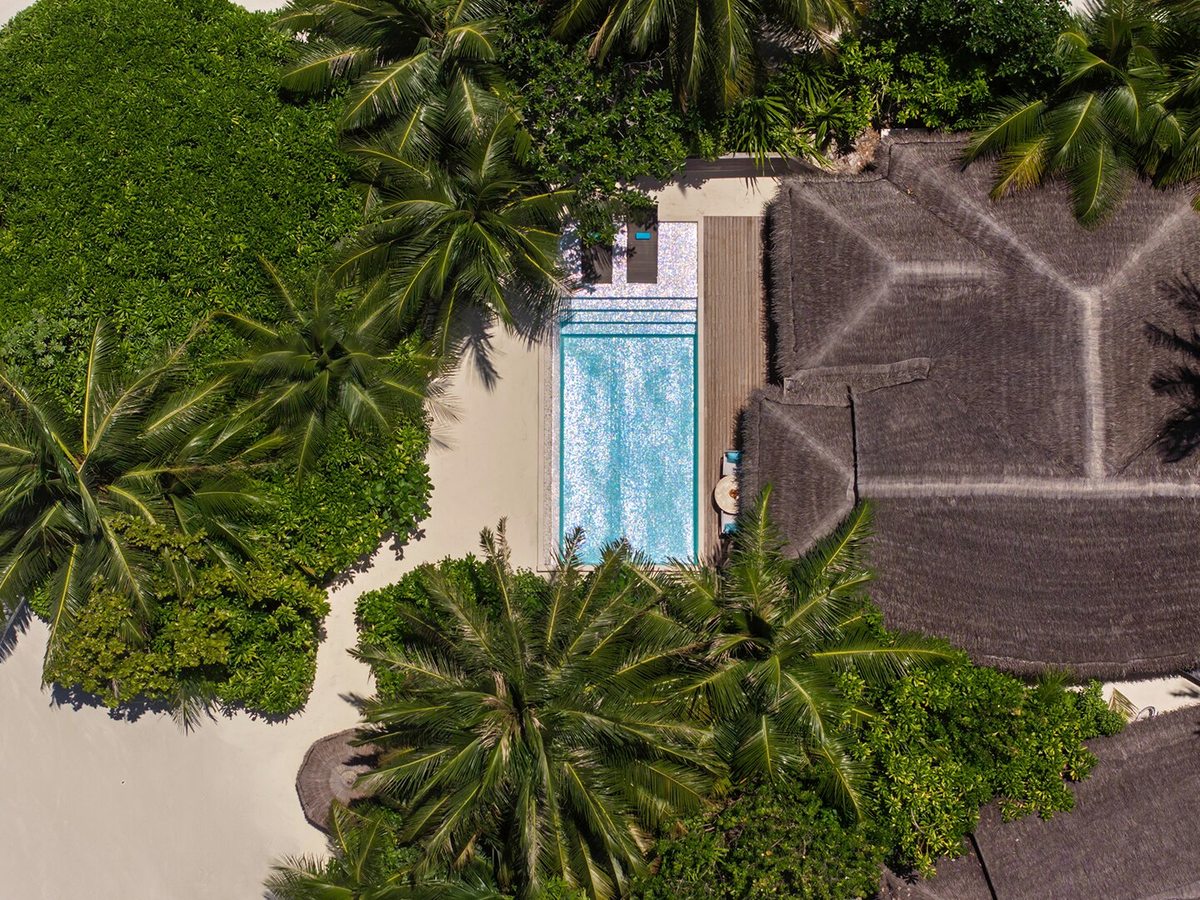 Aerial view of a thatched beach villa with a private pool set among palms right at the edge of a white-sand beach and clear lagoon.