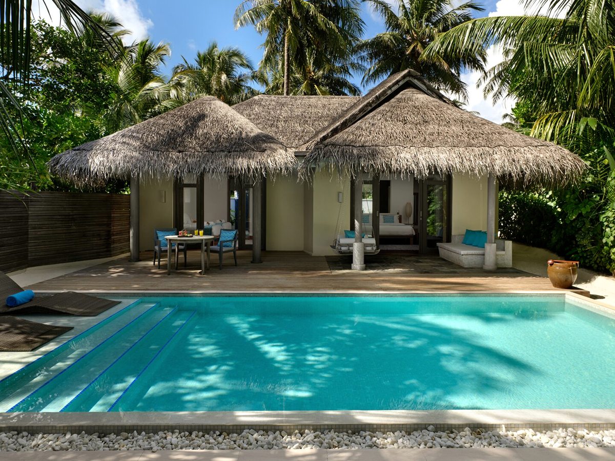 A rectangular private pool on a timber deck beside a thatched villa, with sun loungers, a shaded terrace and tropical planting.
