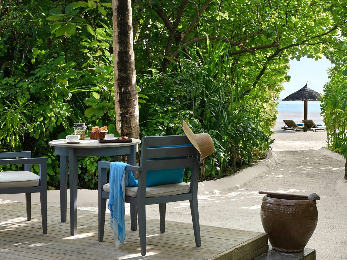 A villa terrace with a round breakfast table and two armchairs on a wooden deck, a shaded sun lounger and a sand path leading to the beach.