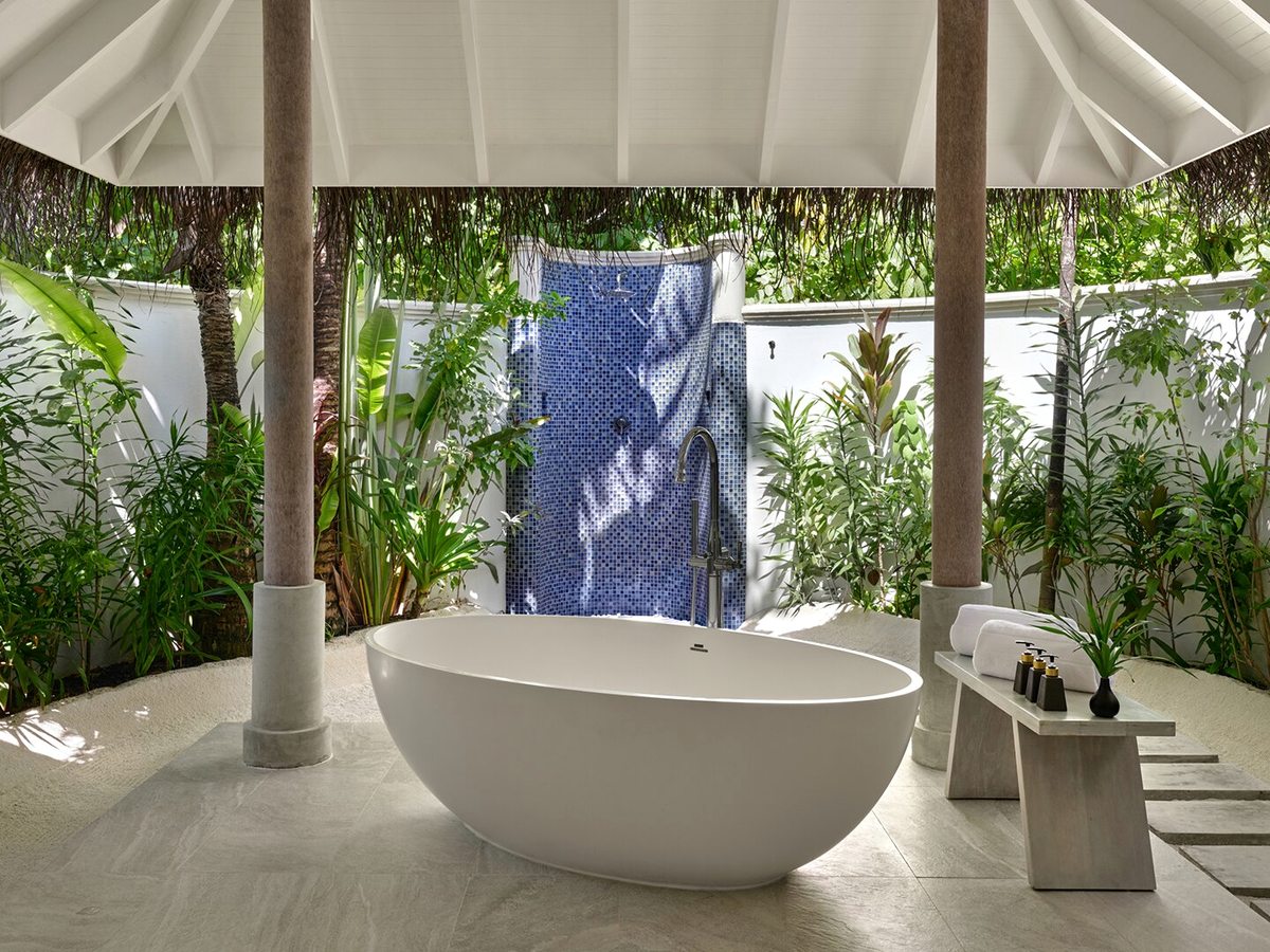 An outdoor bathroom with a freestanding oval bath and a shower against a blue mosaic wall, on stone flooring screened by tropical planting.