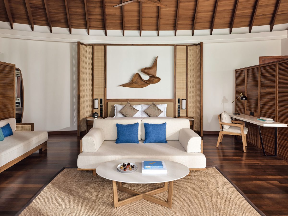 A villa living room with a white sofa and daybed, coffee table, writing desk and television under a vaulted timber ceiling on a polished wood floor.