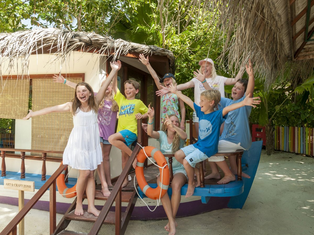 Children with arms raised on a boat-shaped wooden play structure hung with lifebuoys, beside a thatched club building among trees.