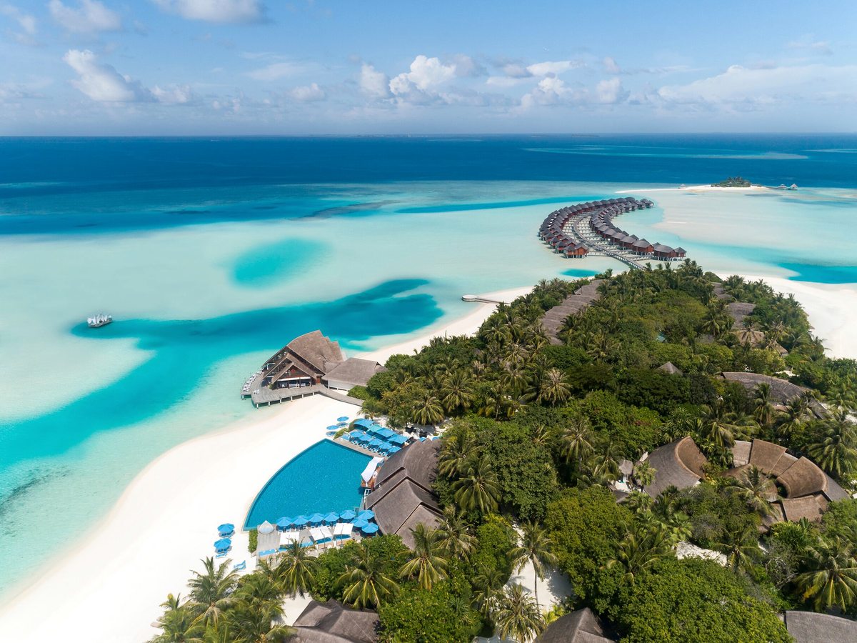 Aerial view of the resort island with a beachfront pool, thatched villas among palms, white-sand beach and a wide turquoise lagoon.