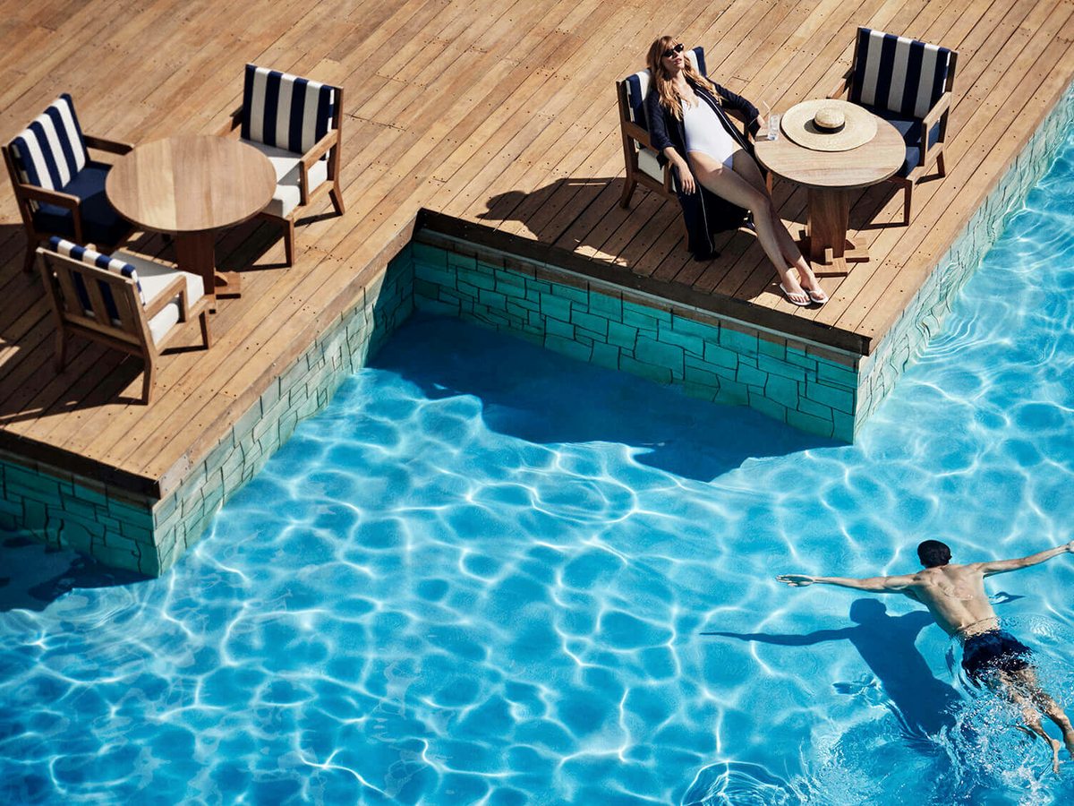 An overhead view of a wooden deck with teak chairs and navy-striped cushions beside a bright blue pool, a woman reclining and a man swimming.