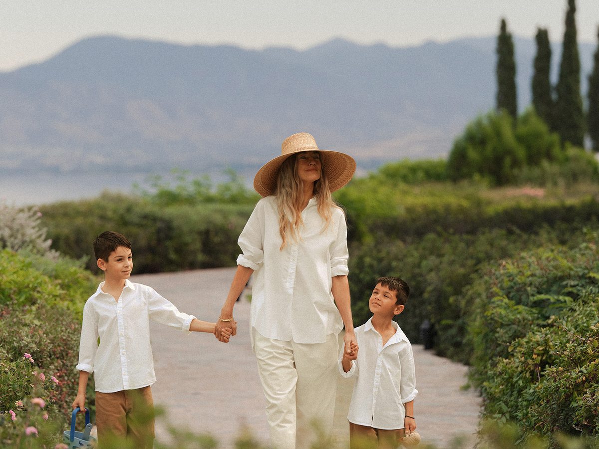 A woman and two boys in white walking hand in hand down a paved garden path edged with flowering shrubs and cypresses, hazy hills and sea behind.
