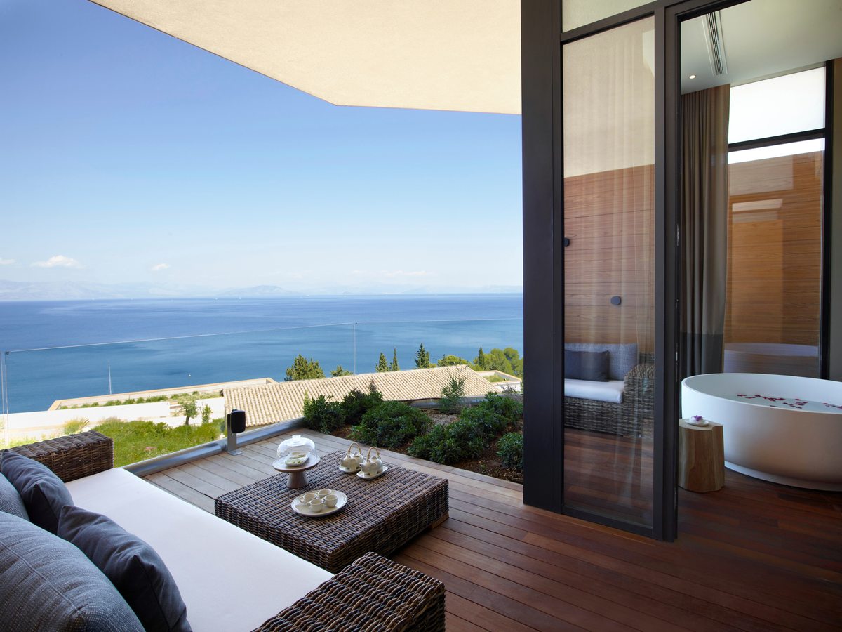 A covered suite terrace with a woven daybed and low table, a freestanding soaking tub just inside the glass doors and a wide sea view.