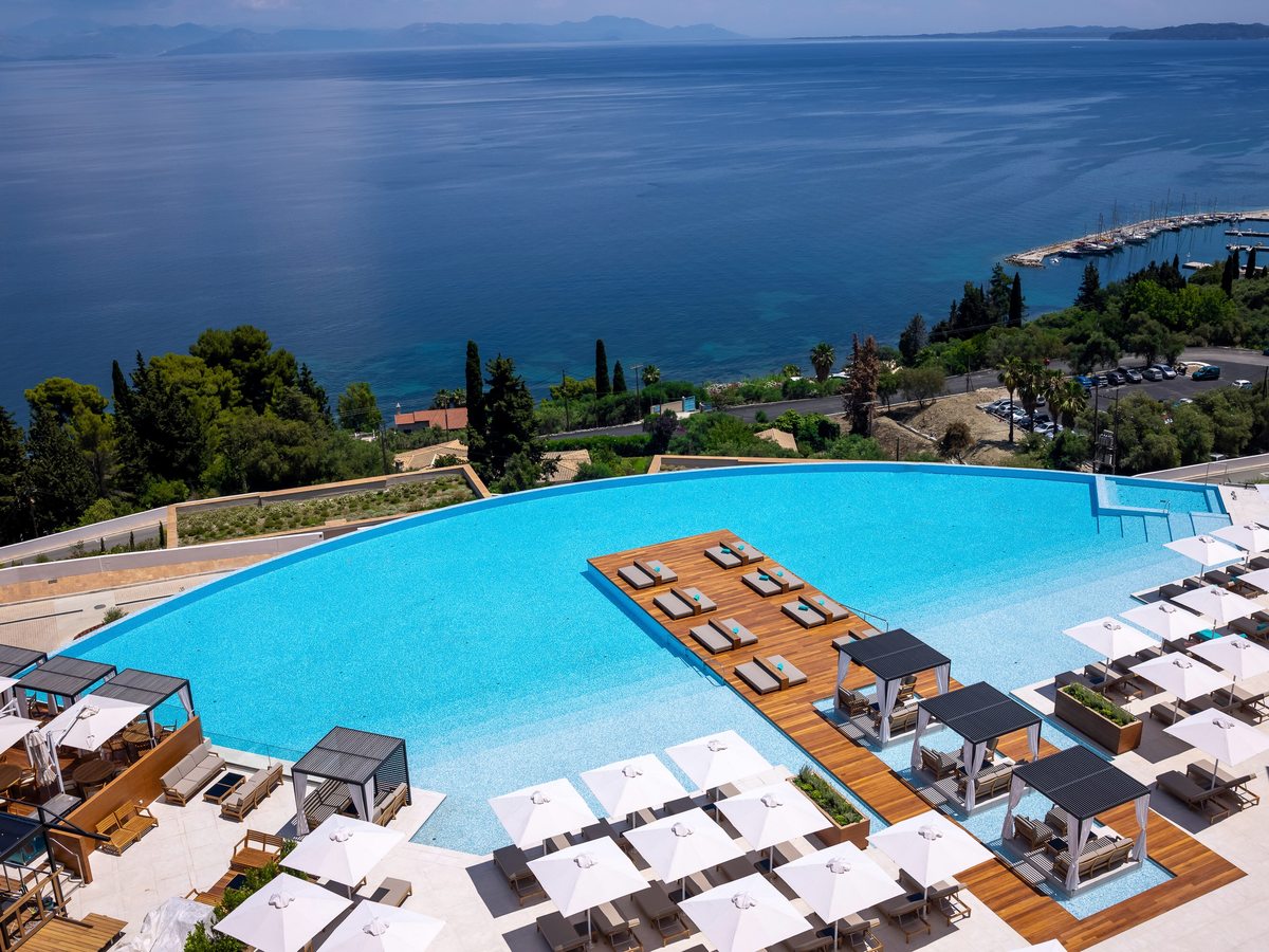 An aerial view of the vast curved infinity pool with a central wooden sun deck, cabanas and rows of white parasols, the deep-blue Ionian and a marina beyond.