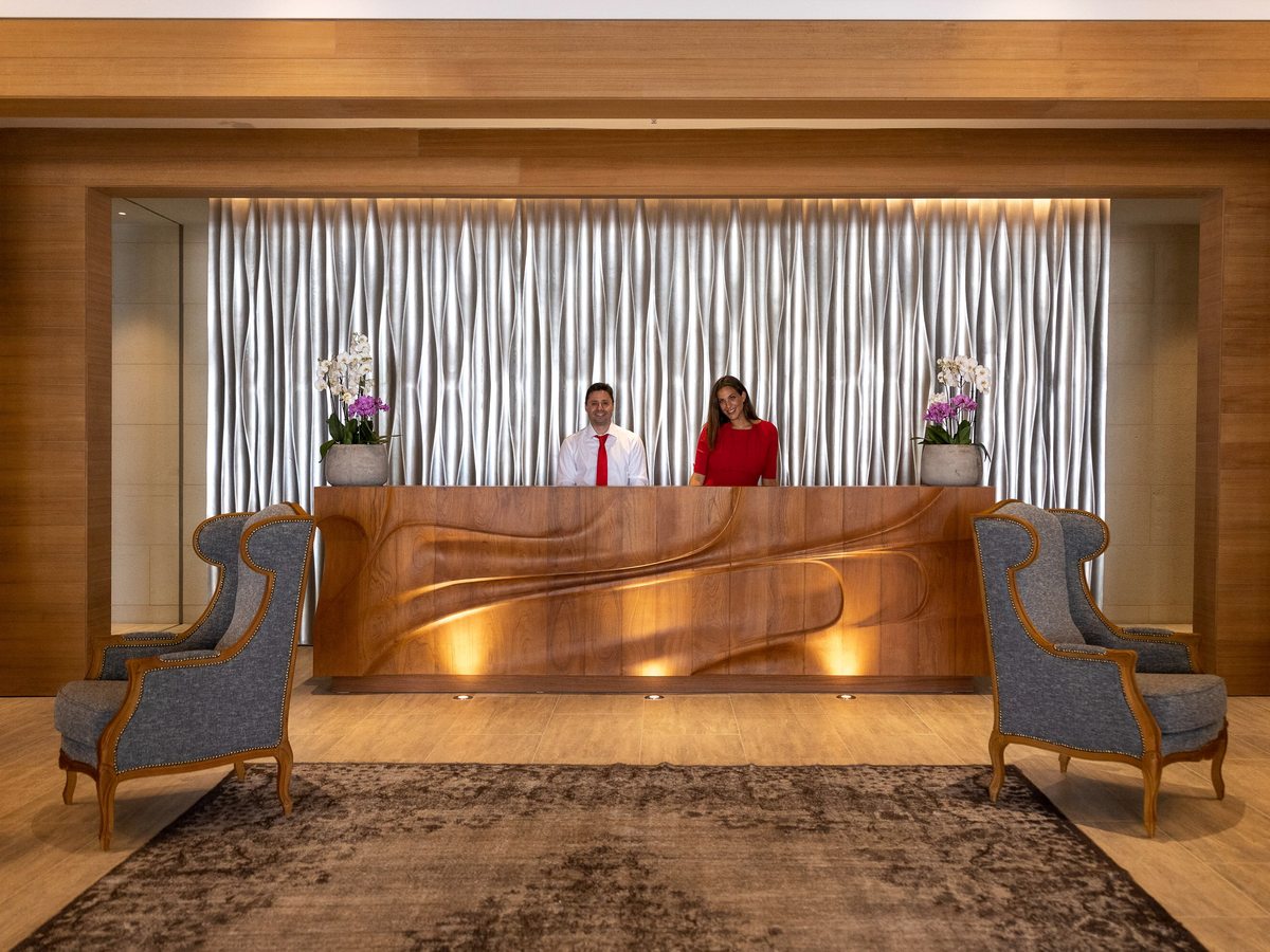 A curved wood-panelled reception desk with two staff behind it, flanked by grey wing armchairs on a patterned rug.
