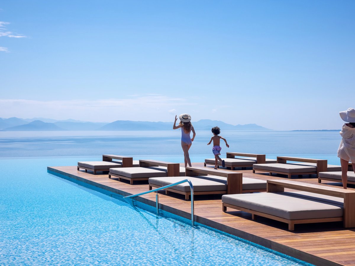 Twin rows of timber sun loungers on a deck that juts into the infinity pool, a woman and a child at the far end above the calm sea and distant mountains.