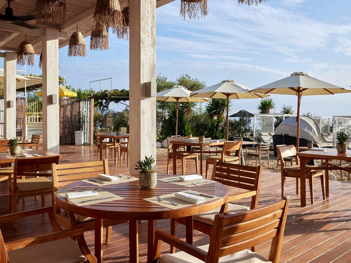A covered beachfront restaurant terrace with round wooden tables and chairs, straw pendant lights and white parasols, open to the sea.