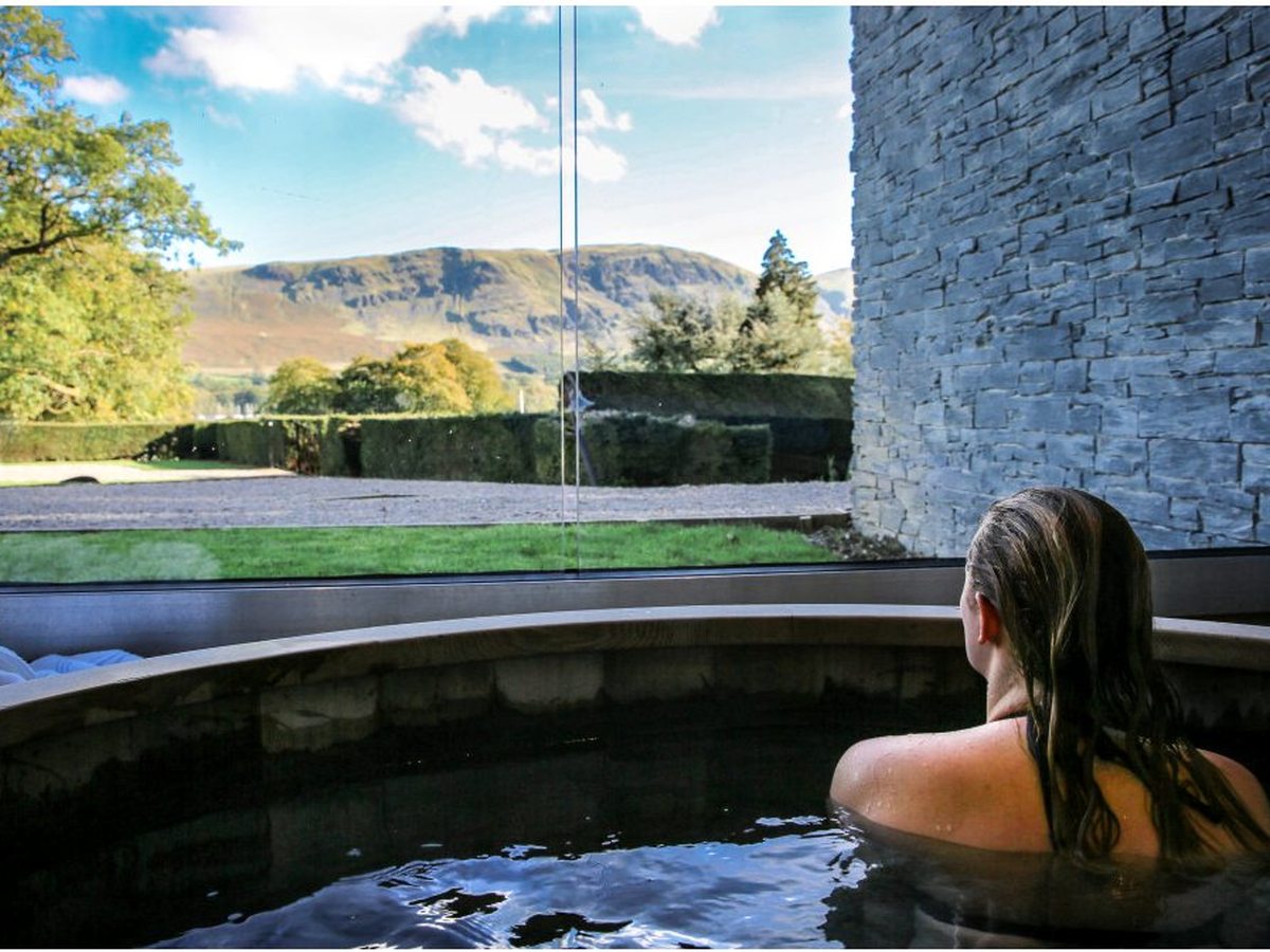 A person in a round wooden hot tub outside, looking across the water to autumn fells.