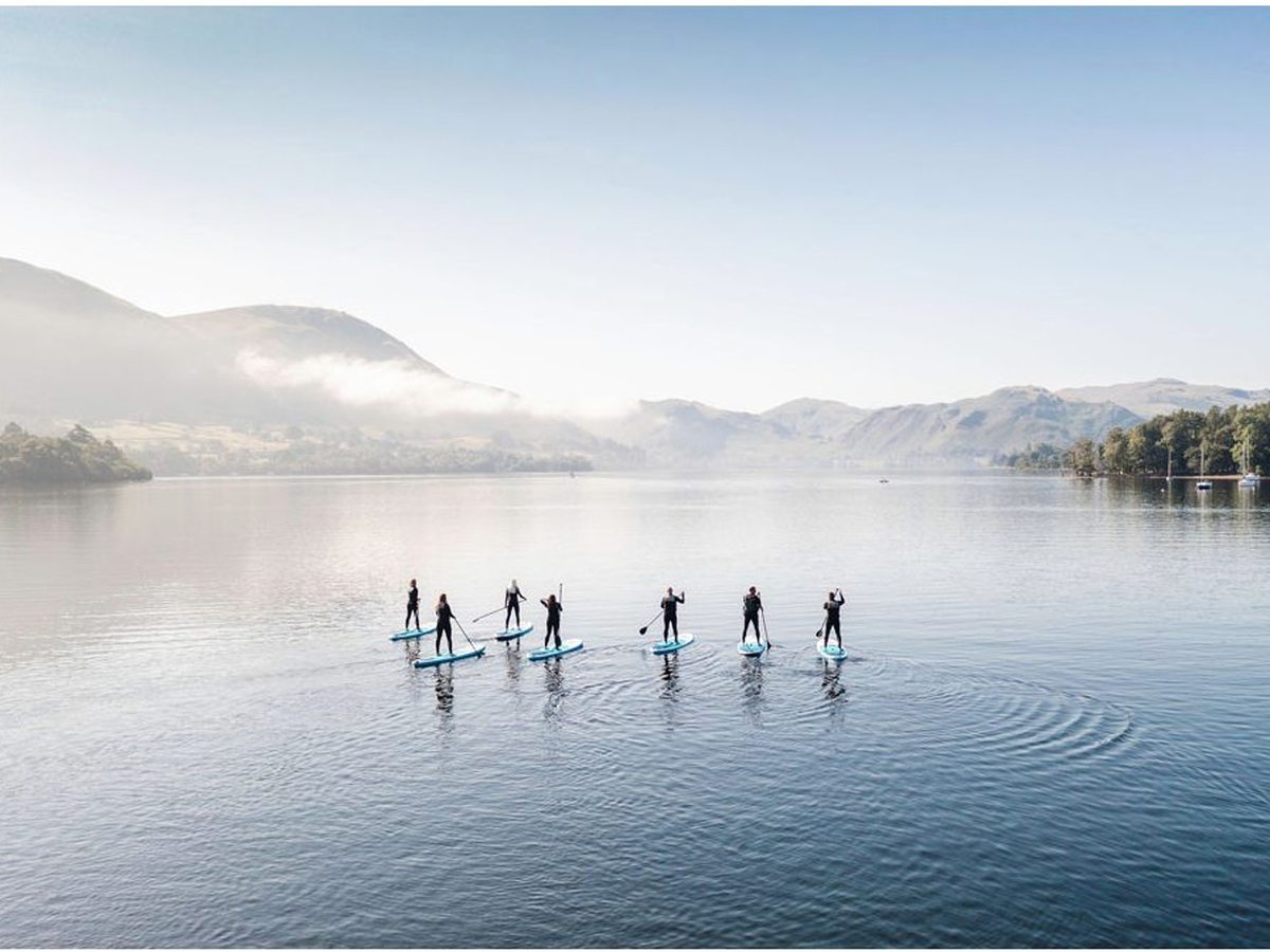 A group of paddleboarders in a line on a still, misty lake with soft fells behind.