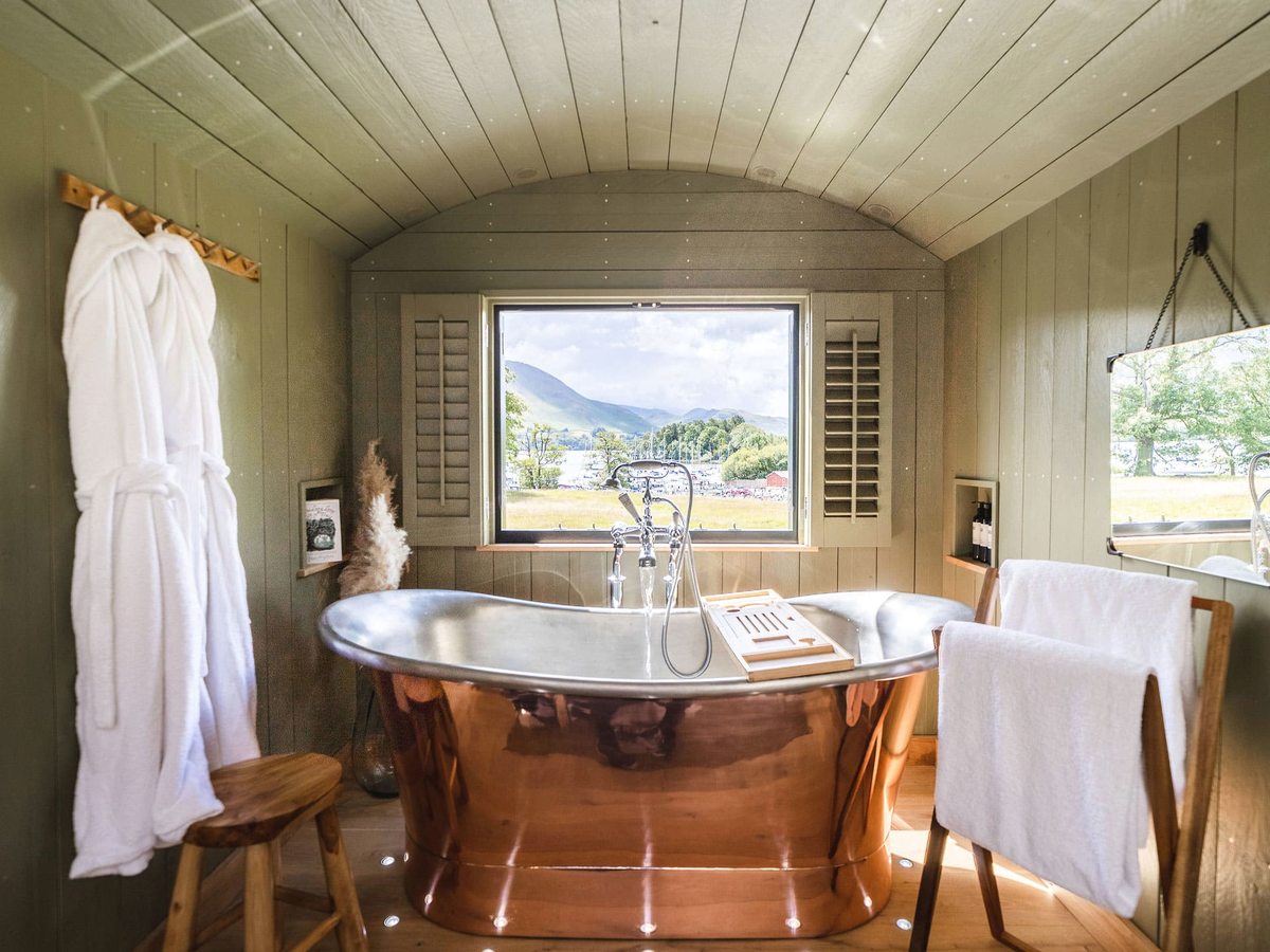 A copper roll-top bath in a sage wood-panelled hut, robes on a peg rail and a window looking to a marina and the fells.