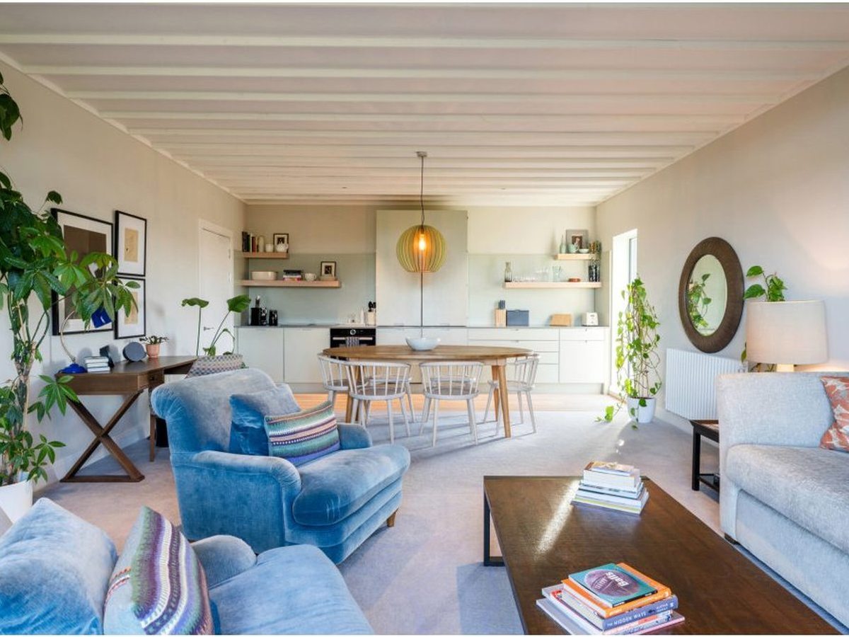 A bright open-plan sitting and kitchen area with pale blue armchairs, a round dining table and a fitted kitchen along one wall.