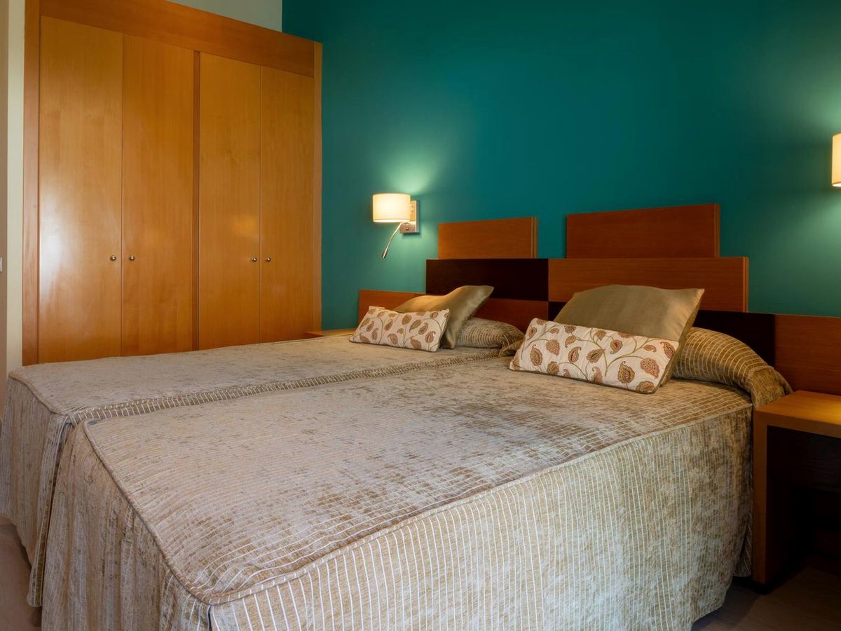Two single beds pushed together with wooden headboards against a teal wall, with fitted wardrobes and wall lamps.