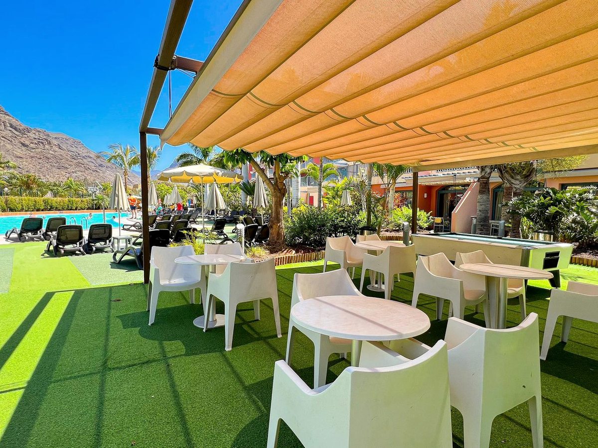 White tables and chairs under a large awning on artificial grass beside the pool, with sun loungers nearby.