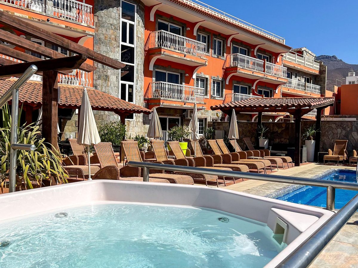 A round outdoor whirlpool tub beside a pool, with sun loungers and an orange apartment building behind.