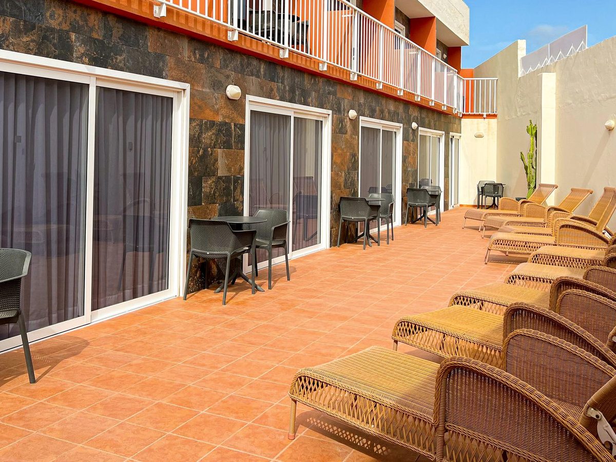 A row of wicker sun loungers on a terracotta-tiled terrace beside sliding doors, below orange apartment balconies.