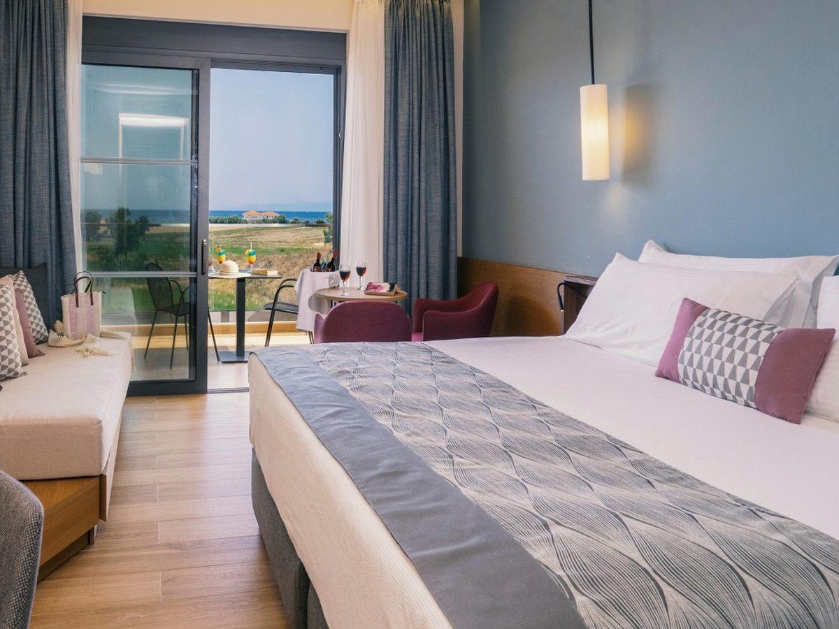 A double room with a large bed, red armchairs and a sofa daybed, sliding doors opening to a private balcony with a coastal view.