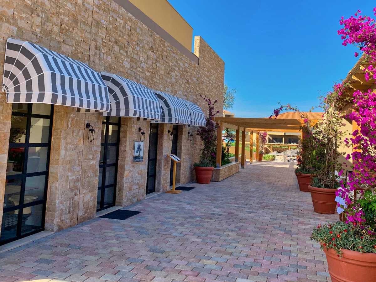 A paved stone walkway between resort buildings, with striped awnings, terracotta pots and climbing pink bougainvillea under a clear blue sky.