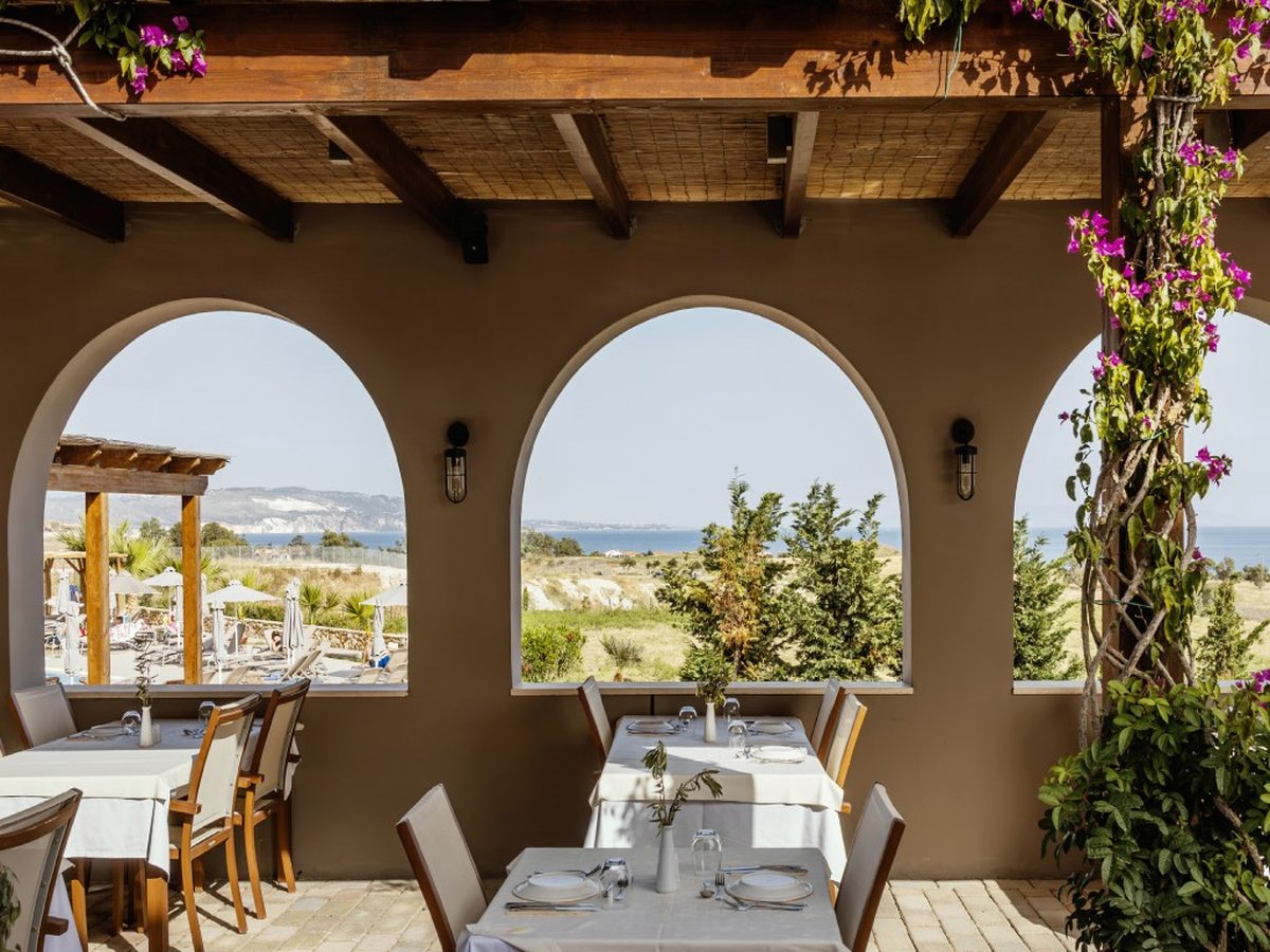 A covered dining terrace with arched openings framing the countryside and sea, white-clothed tables and climbing bougainvillea.