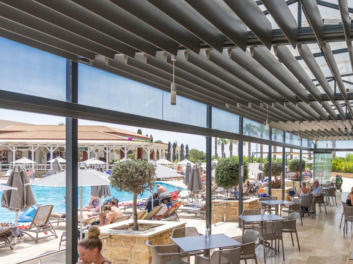A covered terrace with tables and chairs looking onto the pool, sun loungers and parasols, with a grill bar behind.