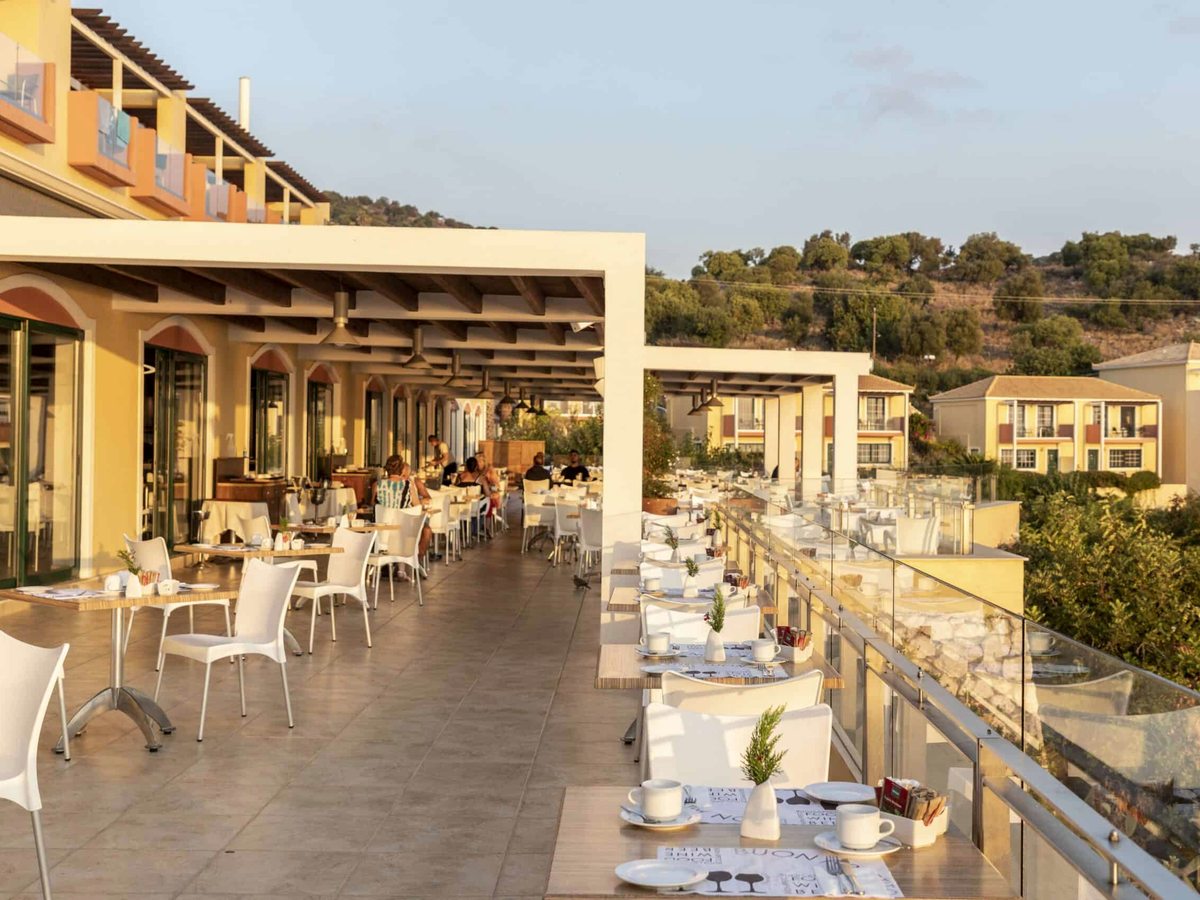 Rows of laid tables and white chairs on a long paved terrace under a pergola, with the hillside behind.