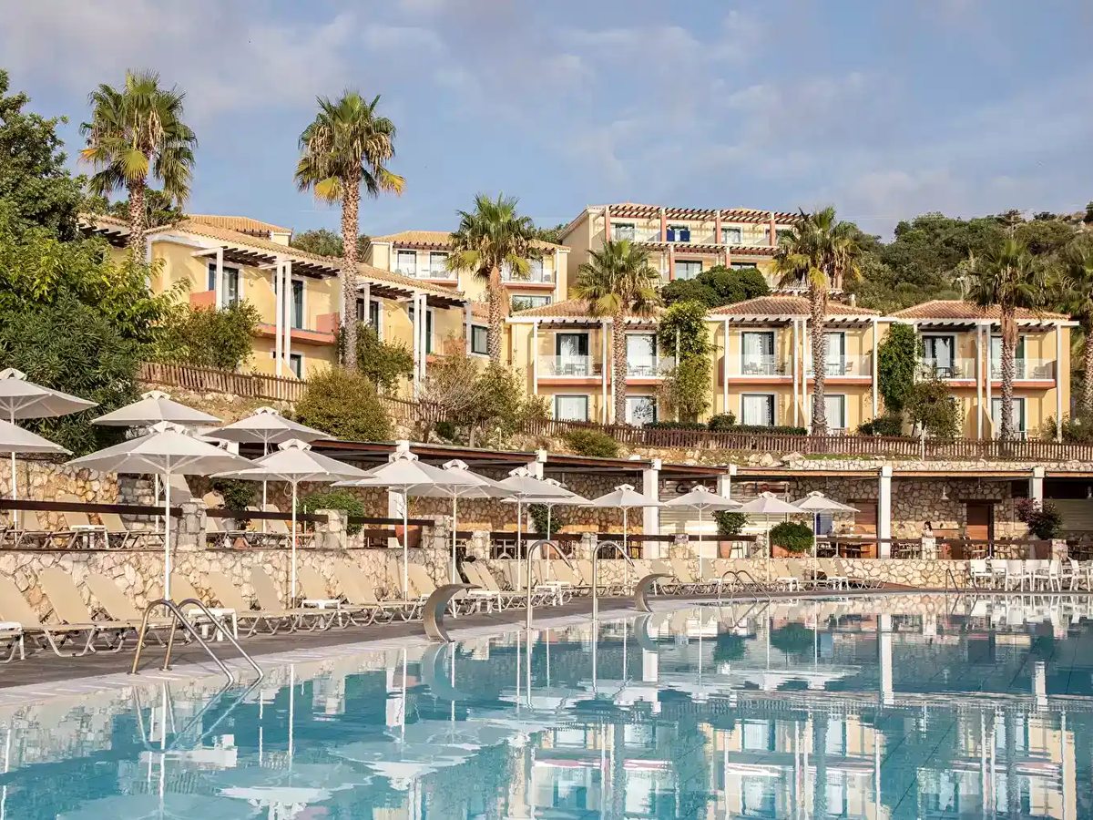 A large irregular pool with sunbeds and white parasols on a paved deck, palm trees and terraced buildings on the hillside behind.