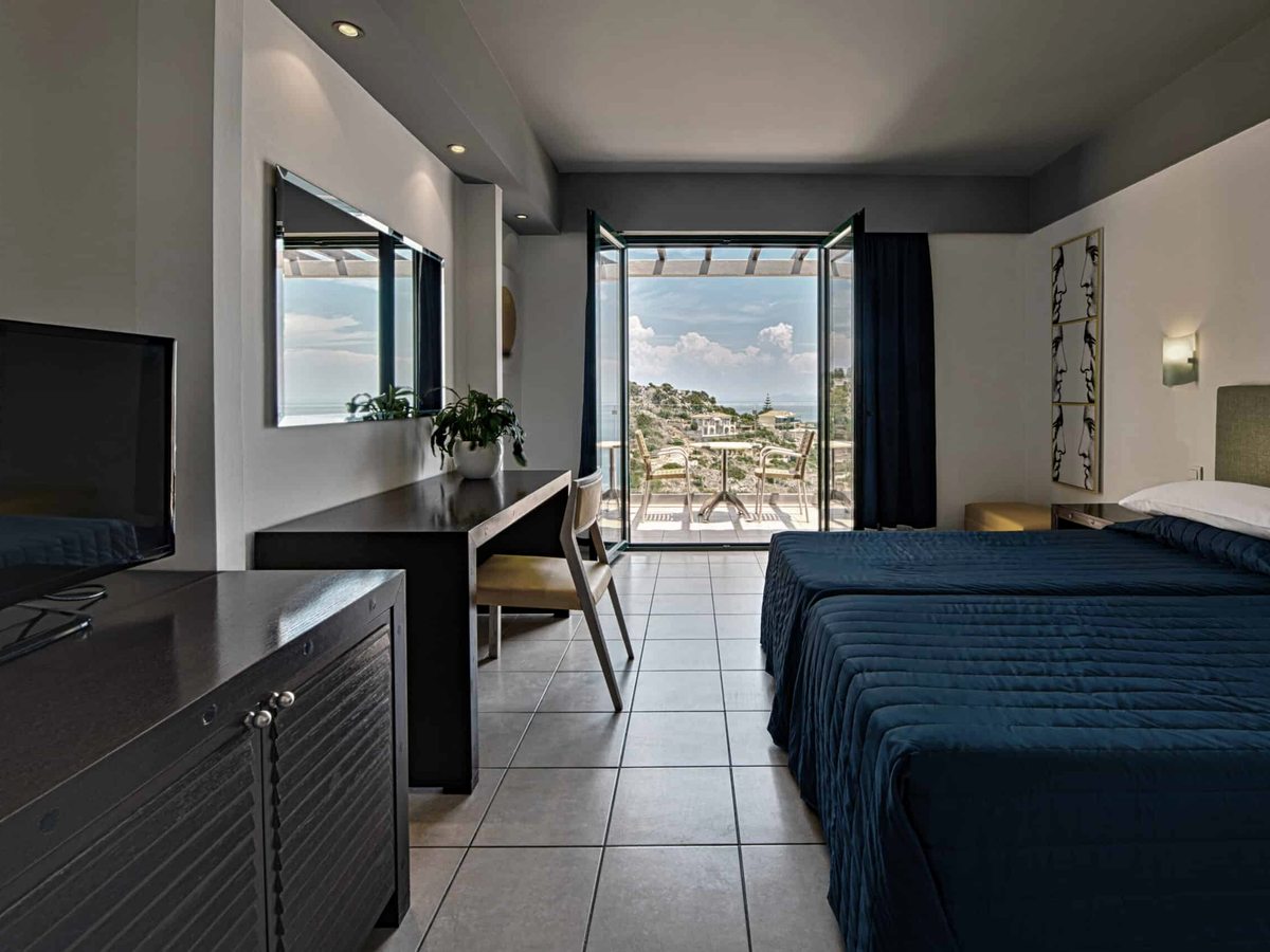 A double room with a blue bedspread, a desk and a wall-mounted TV, with balcony doors to a sea view.