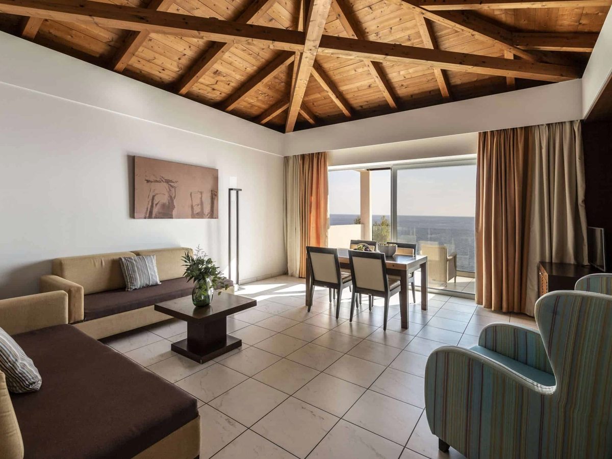A suite living room with two sofas, a dining table and an armchair under a pitched timber-beamed ceiling, with sliding doors to a sea view.