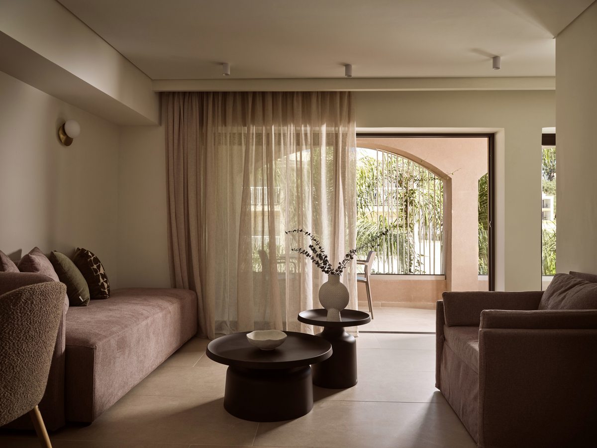 A living area with a sofa and armchair, round coffee tables and sliding doors opening onto a covered terrace with sheer curtains.
