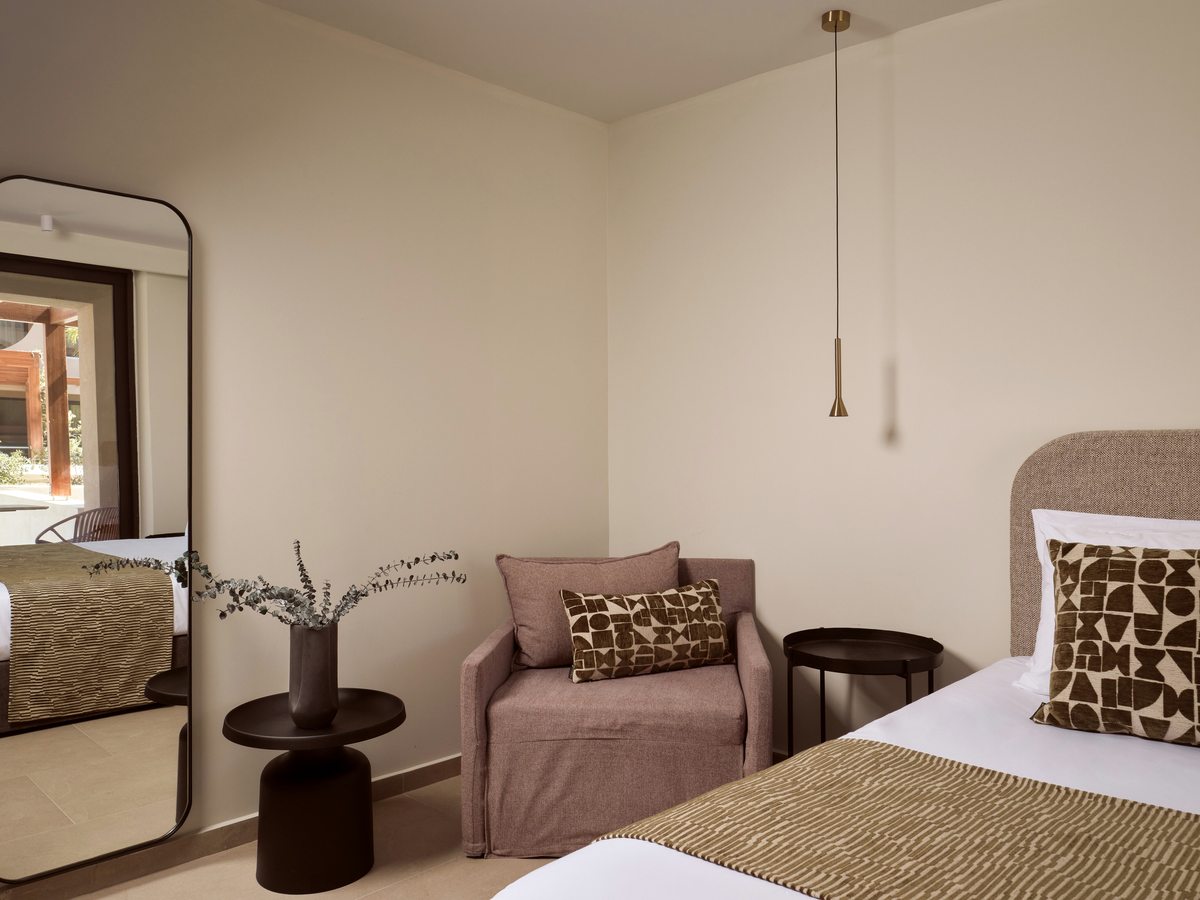 A bedroom with white bedding and a green runner, a compact pink sofa, a round side table, a tall floor mirror and a pendant light against beige walls.