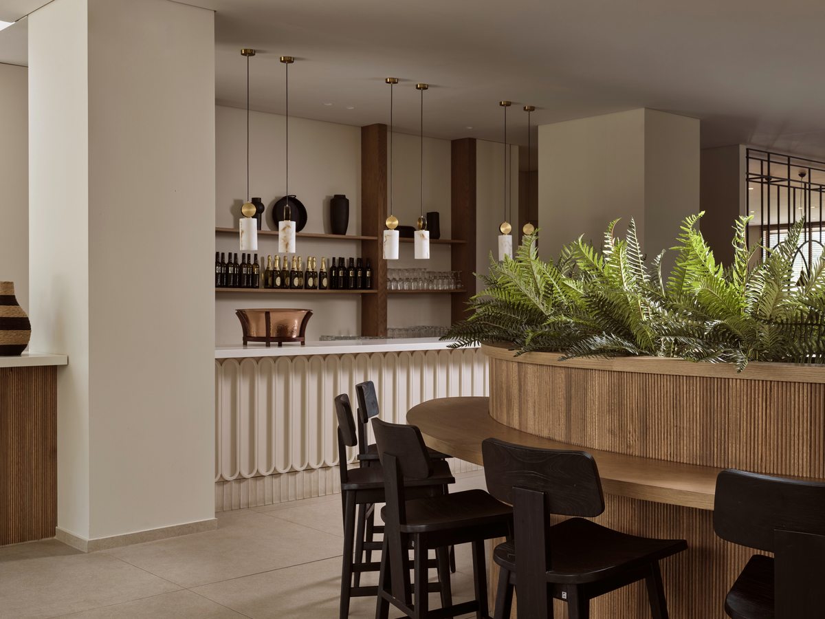 An indoor bar with a ribbed timber counter, bottle shelves behind and high stools, screened from the dining tables by a tall fern planter.