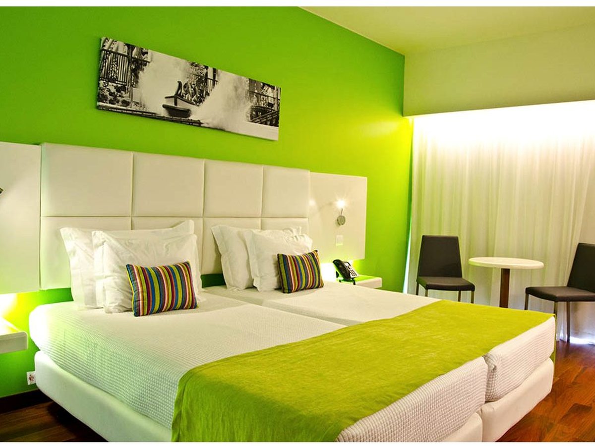 A twin room with two single beds against a green feature wall, a padded white headboard, a small table and chairs and wood flooring.