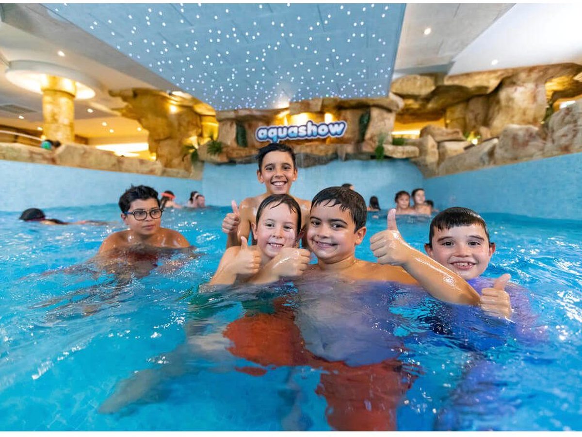 Children smiling and giving thumbs up in a blue indoor pool with rock-effect walls and a starlit ceiling.