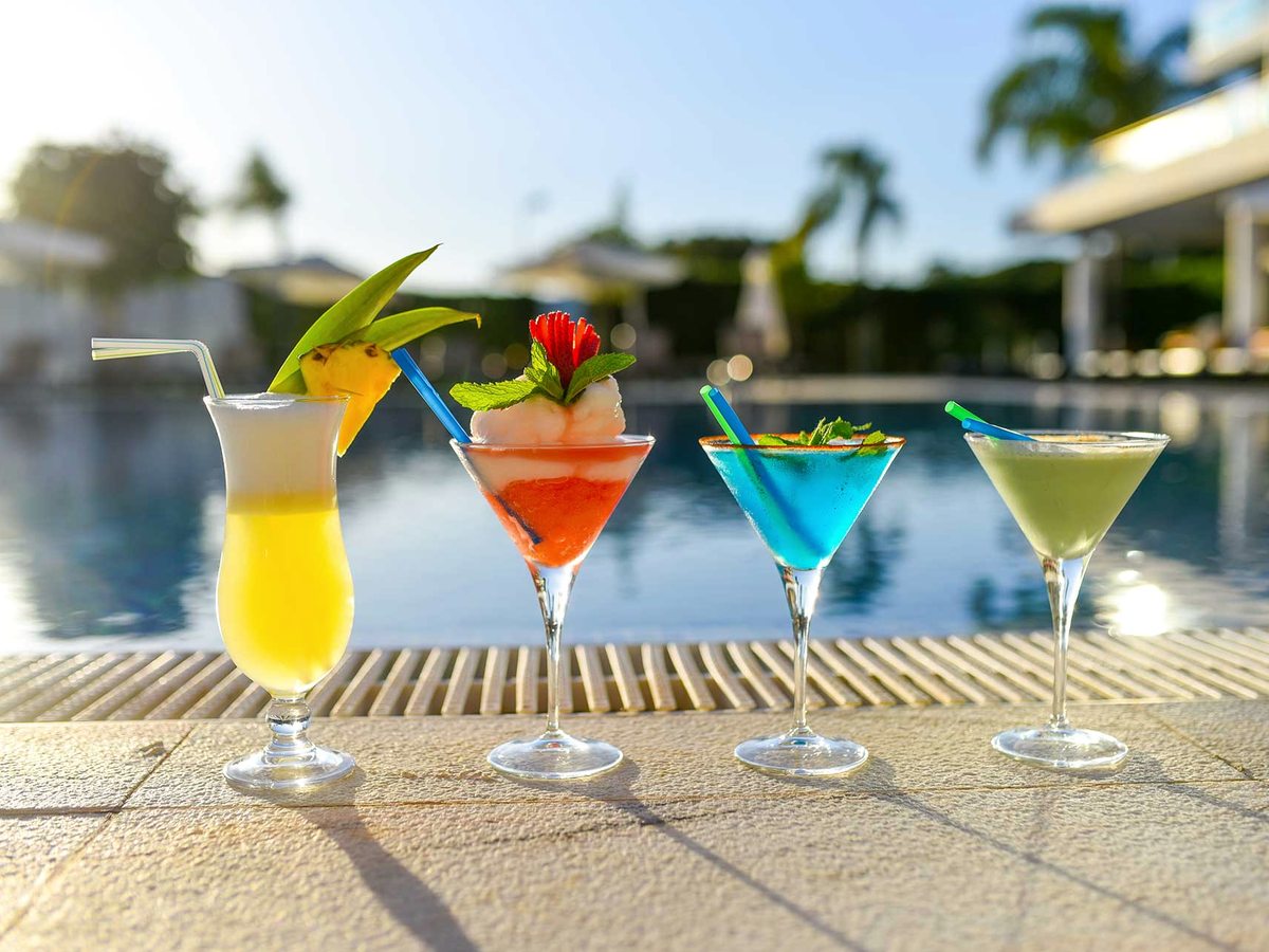 A row of colourful cocktails on the tiled edge of the outdoor pool with parasols and palms behind.