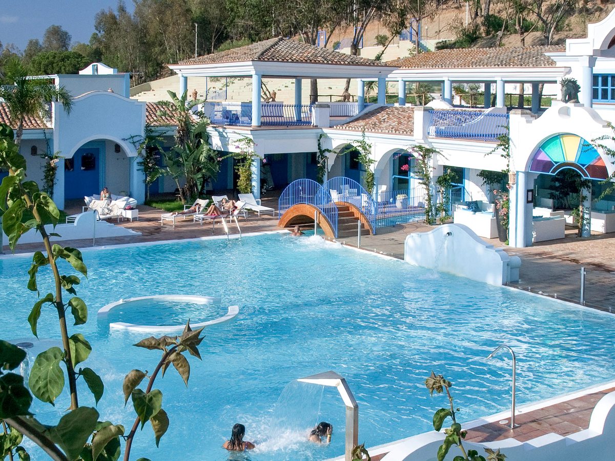 A multi-level pool with a small arched footbridge, set among white buildings with blue trim, arcades and a stained-glass fan window.