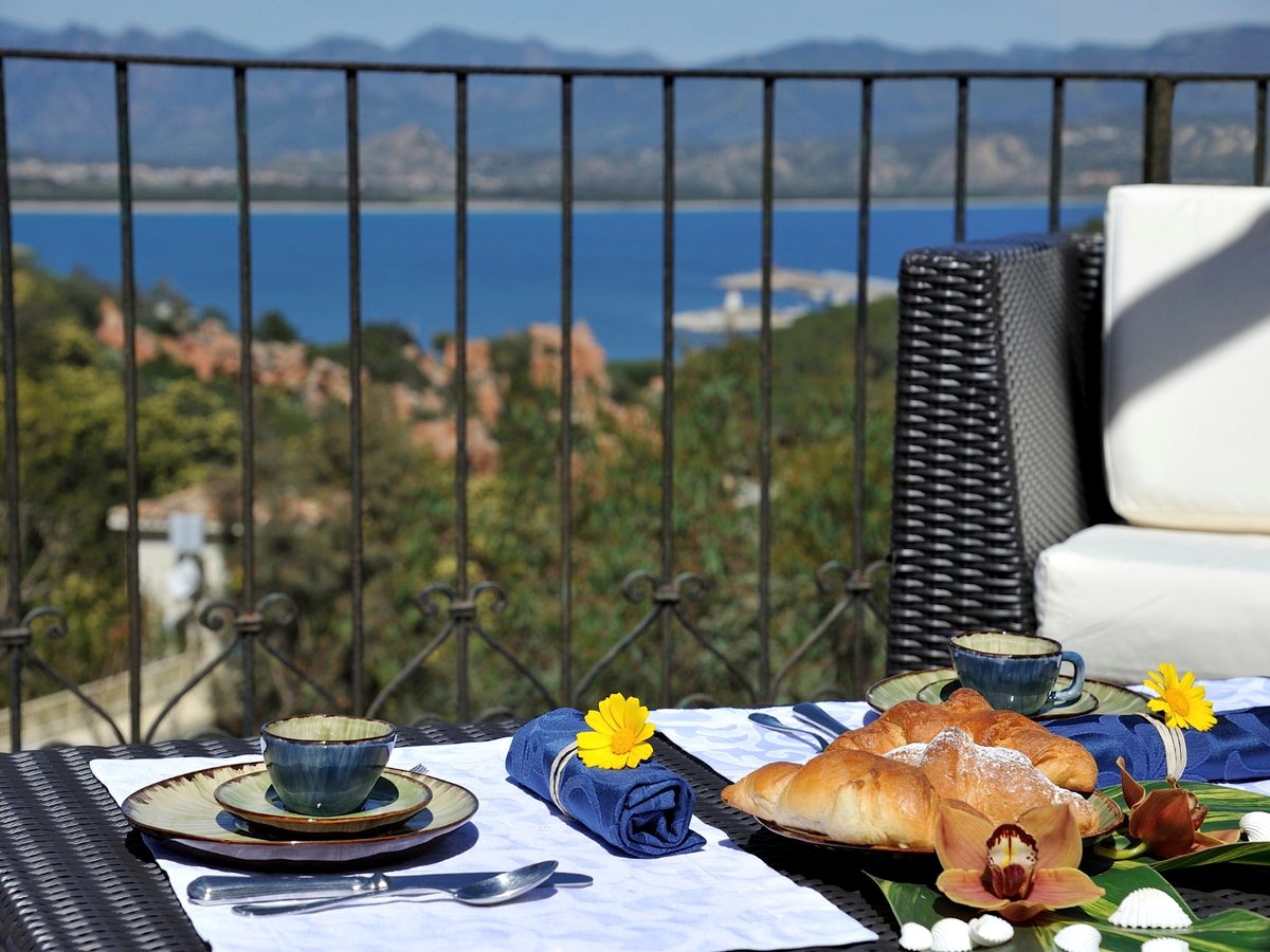 A breakfast of croissants, coffee in glazed cups and an orchid set on a terrace table, with the bay and red rocks through the railing behind.