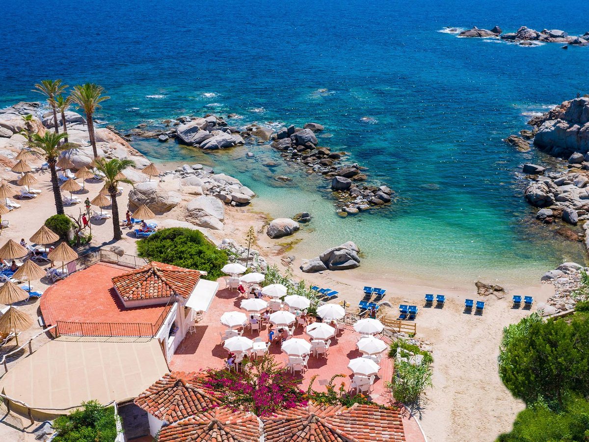 Aerial view of a cove with a terracotta-tiled beach-bar terrace, rows of thatched and blue loungers on pale sand and clear water over rocks.